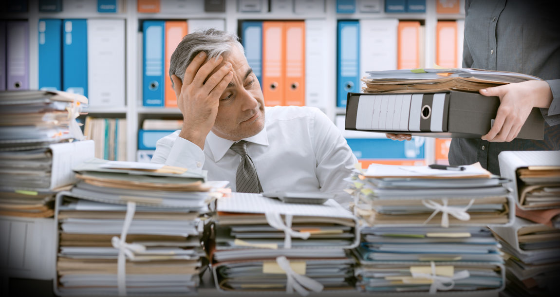 Image of man looking bored and exhausted seated in front of large stacks of documents.