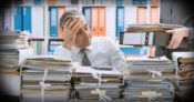Image of man looking bored and exhausted seated in front of large stacks of documents.