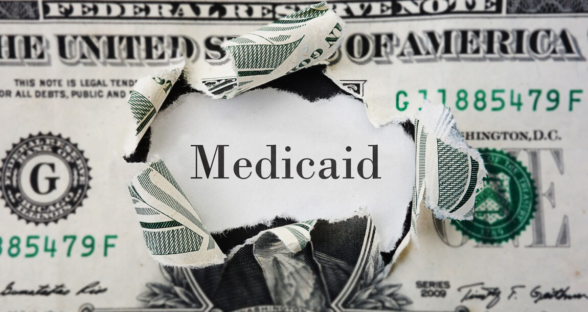 Image of the word "Medicaid" under a hole ripped in a dollar bill