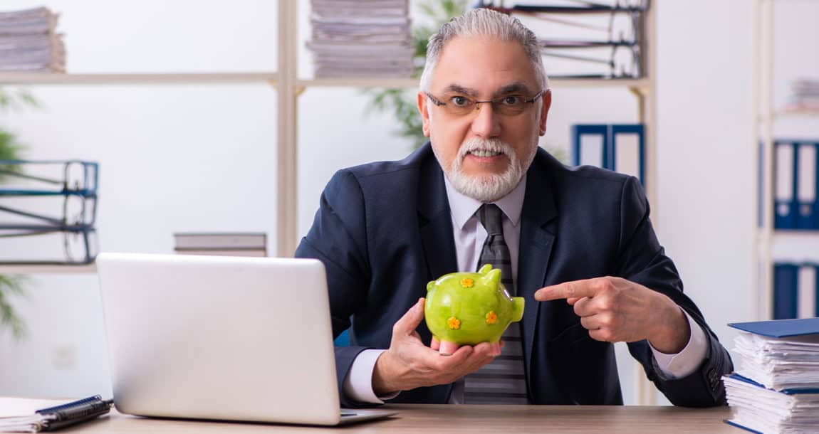 Image of man in suit and tie pointing to a piggy bank he's holding, while sitting at a desk with an open laptop.