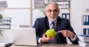 Image of man in suit and tie pointing to a piggy bank he's holding, while sitting at a desk with an open laptop.