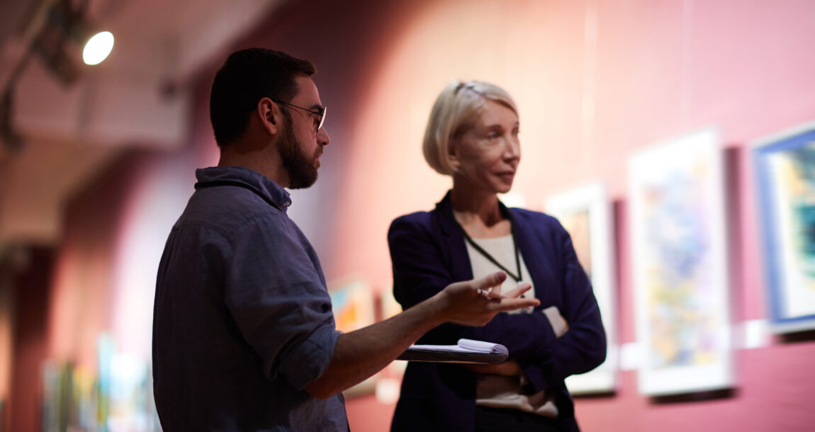 Image of professionally dressed man and woman talking while standing in front of several paintings.