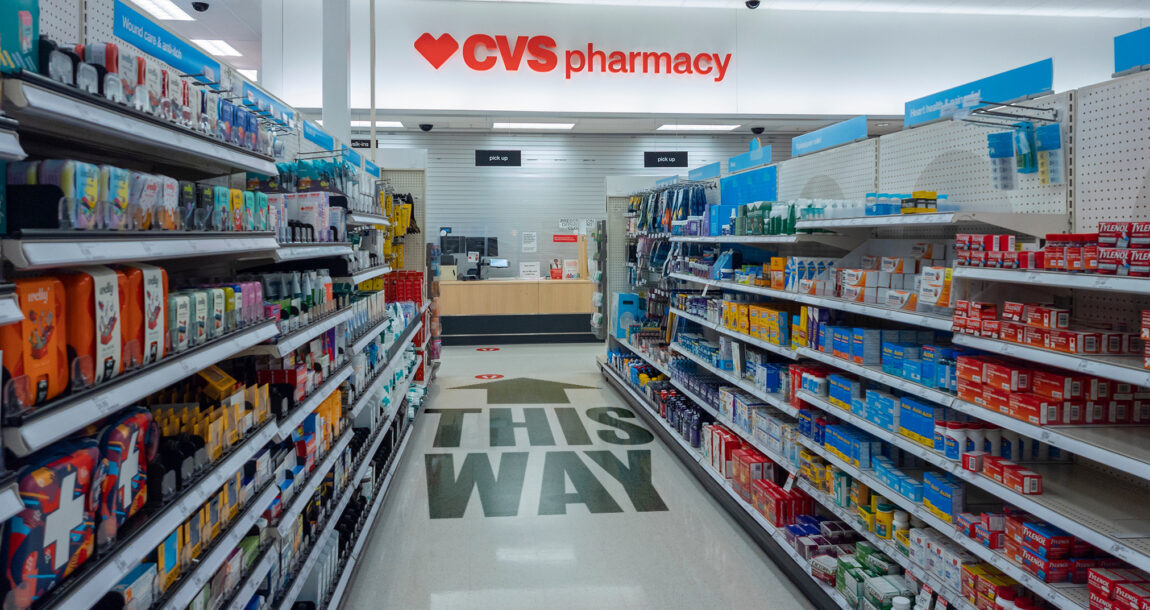 Image of aisle in CVS leading to the Pharmacy with the phrase: This Way. Oklahoma Dept. of Insurance accuses CVS of steering customers to its own pharmacy.
