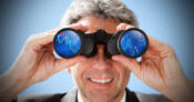 Picture shows a man looking through binoculars with index results reflected in the lenses.
