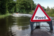 Image of a flooded road with a sign that reads "Flood" in the foreground.