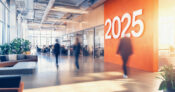 Photo illustration of a workplace with a large "2025" on the wall. Emerging-workplace-benefit-trends-for-2025.